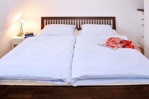 Ein Doppelbett mit weißen Bettwäsche, ein hölzerner Kopfteil, ein Nachttisch mit Lampe und eine rote, gemusterte Decke.