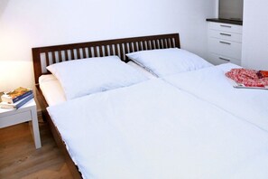 A double bed with white bedding, a wooden headboard, a bedside table with books, and a small drawer unit. - House Hahnen - Ground Floor (Norderney)