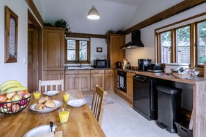A kitchen with wooden cabinets, a dining table set with fruits and breakfast items, and a black dishwasher. - Willow Tree Barn (Bideford)