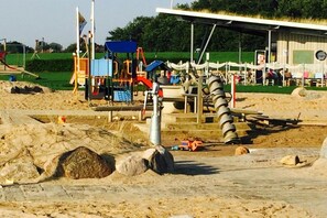 Une aire de jeux dotée de diverses structures ludiques, d’un espace de repos couvert et d’un sol sablonneux.