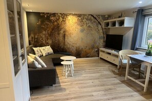 Un salon moderne comprenant un canapé, deux tabourets blancs, une table à manger avec une chaise, un téléviseur et un mur décoratif.