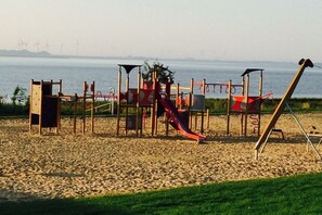 A playground with a slide and climbing structures, located on a sandy area with a body of water and wind turbines in the background. - Holiday apartment with 1 bedroom (Tönning)