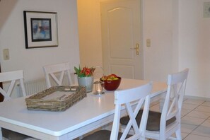 A dining area with a white table, two white chairs, a framed picture on the wall, a vase with flowers, and a bowl of fruit. - Möhring Holiday Farm (Warwerort)