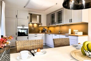 A modern kitchen with white cabinets, a dining table with wicker chairs, and a breakfast setting. - Holiday apartment with 2 bedrooms (St. Peter-Ording)