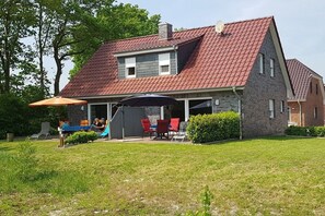 A two-story house with a red-tiled roof, a covered patio area with outdoor furniture, and a small play structure. - Holiday apartment with 2 bedrooms (Aurich\/ Tannenhausen)