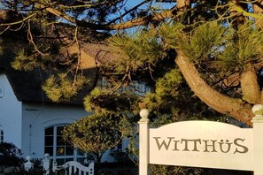 A sign with the name 'WITHUS' in front of a building with a thatched roof and pine trees. - Witthüs holiday apartment 16 (Wenningstedt)