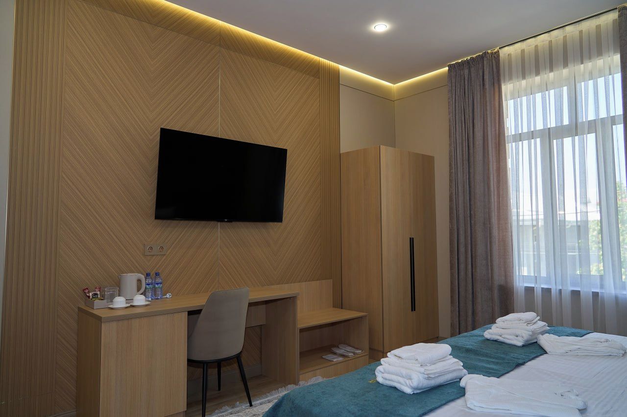 Habitación de hotel con cama, escritorio, silla, televisor montado en la pared y ventana con cortinas.