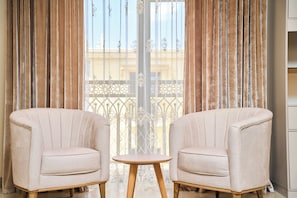 Una habitación con dos sillones mirando a una pequeña mesa redonda, un balcón con barandilla de hierro forjado y vistas a un edificio a través de la ventana.
