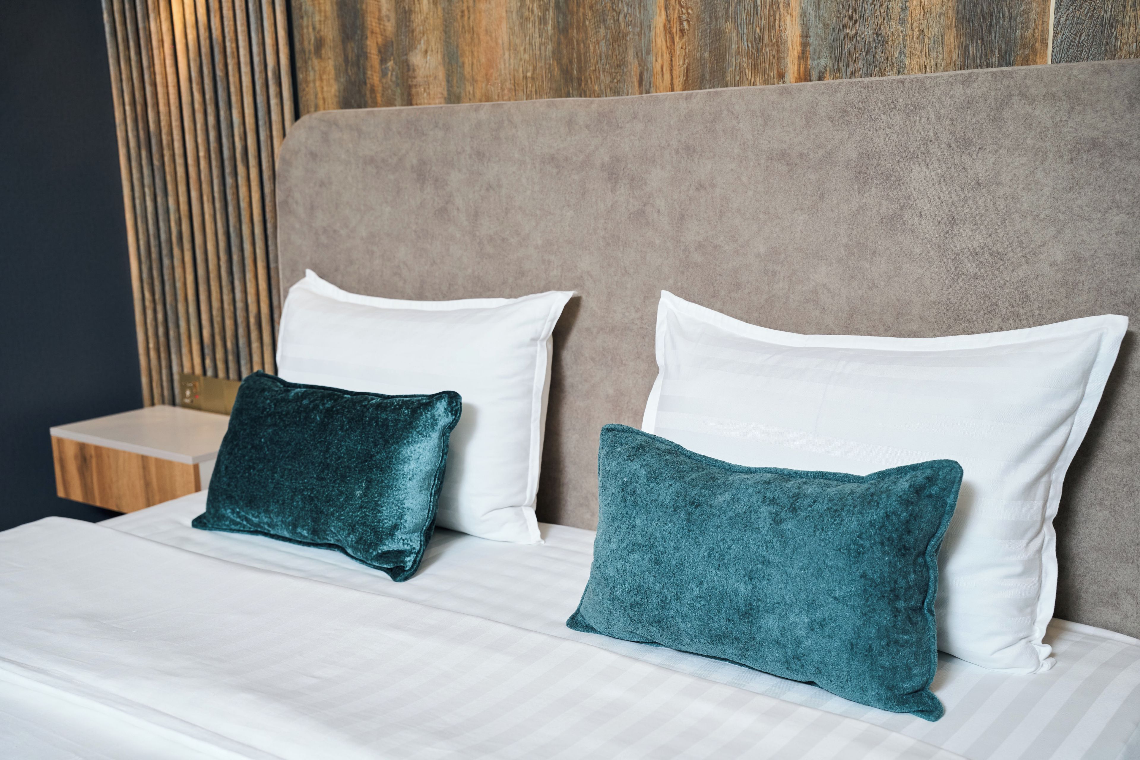 A bed with two white pillows and two teal pillows, a wooden headboard, and a nightstand with a lamp.