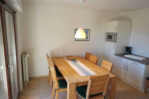 A dining area with a wooden table and chairs, a hanging light fixture, and a kitchen area with cabinets and a countertop. - Guesthouse "Wangerland" (Horumersiel)