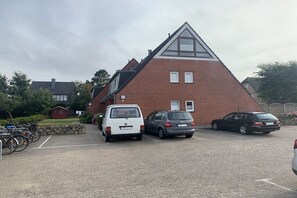 A parking area with three cars and bicycles, a red brick building with a triangular roof, and a stone wall. - Holiday apartment in Sylt with 1 bedroom (Wenningstedt)