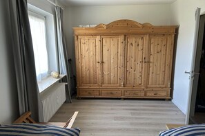 A room with a wooden wardrobe, a radiator, a window with curtains, and a bed with striped bedding. - Holiday apartment in Sylt with 1 bedroom (Wenningstedt)
