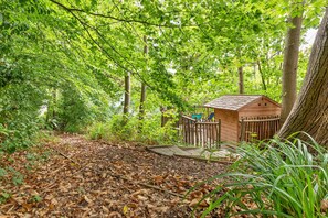 A wooden structure with a sloped roof, surrounded by dense foliage and a ground covered with dry leaves. - The Blue Barn (Tunbridge Wells)