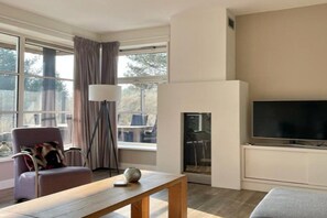 Ein modernes Wohnzimmer mit Flachbildfernseher, Kamin, bequemen Sitzgelegenheiten und großen Fenstern.