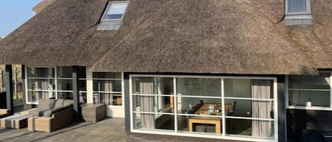 Et stråtækt hus med et stort glasvindue, udendørs siddepladser og et synligt interiør.