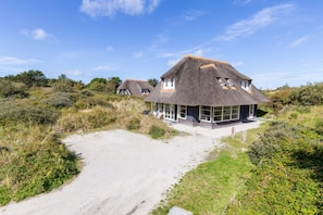 A thatched-roof house with large windows, surrounded by natural vegetation and a sandy path. - Luxury dune villa on Ameland (Nes)