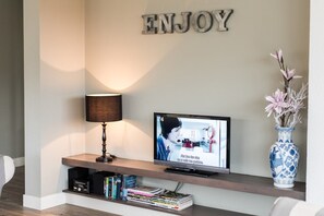 Een flatscreen televisie op een houten plank, een lamp, een vaas met bloemen, en een bord met de tekst 'ENJOY' aan de muur.