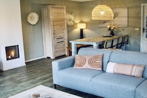 A living room with a sofa, a fireplace, a dining table, and a pendant light. - Charming villa in the dunes of Ameland (Nes)