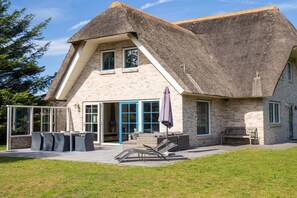 Ein Haus mit einem Strohdach, großen Fenstern und einer Terrasse mit Gartenmöbeln.