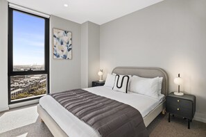 A modern bedroom with a large bed, two bedside tables, a nightstand, a window with a city view, and a wall-mounted abstract painting. - Skyline 3-bed Apartment Near South Melbourne (Melbourne)
