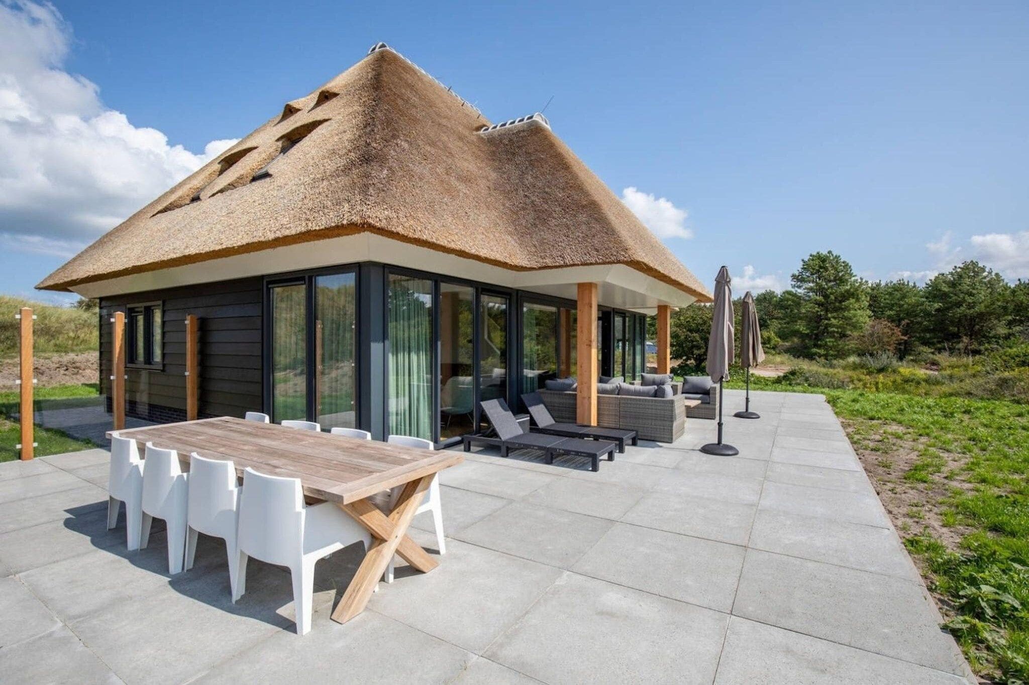 A modern house with a thatched roof, large windows, a dining area with a wooden table and white chairs, and outdoor seating with umbrellas.