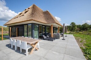 Ein modernes Haus mit einem Strohdach, großen Fenstern, einem Essbereich mit einem Holztisch und weißen Stühlen sowie Sitzmöglichkeiten im Freien mit Sonnenschirmen.