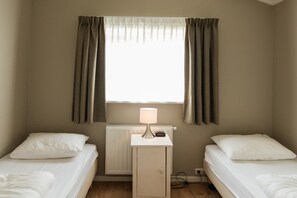 A small, single-room accommodation with two beds, a bedside table with a lamp, and a window with curtains. - Charming bungalow on Ameland with a beautiful garden (Ballum)