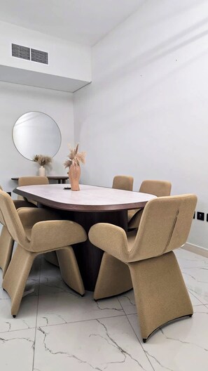 Een moderne eetruimte met een ronde tafel, zes stoelen, een vaas met gedroogde planten, een ronde spiegel en een witte muur met een ventilatie rooster.