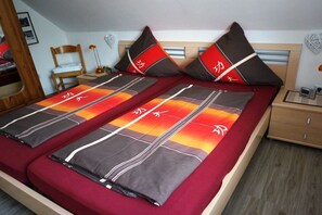 A double bed with a red and orange patterned bedspread, two pillows with red and black designs, a wooden headboard, a bedside table with a lamp, and a small bedside table with a book and a remote control. - Baltrum at the "Kogge" house (Norden-Norddeich)