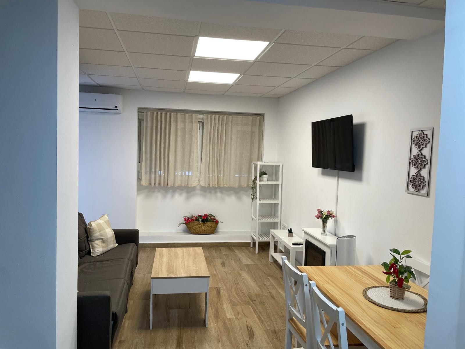 Una sala de estar amoblada con un sofá, una mesa de centro, un televisor montado en la pared y un ventanal con cortinas.
