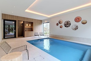 A modern indoor swimming pool with a clear blue water surface, surrounded by lounge chairs and a wall with circular decorative elements. - Family retreat in Belgium with sauna (Rochefort)