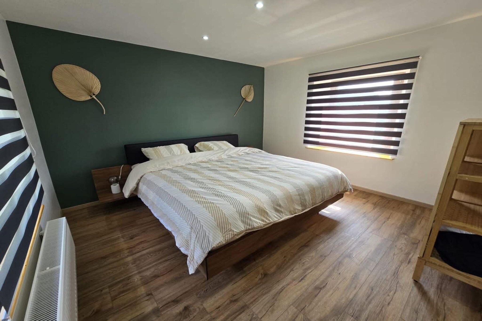 Een slaapkamer met een bed, houten vloeren, een raam met zonwering en een ventilator aan de muur.