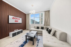 Ein modernes Wohnzimmer mit einem großen Fernseher, der 'BOOK ME' anzeigt, einem Sofa, einem Couchtisch und Blick auf Gebäude durch ein großes Fenster.