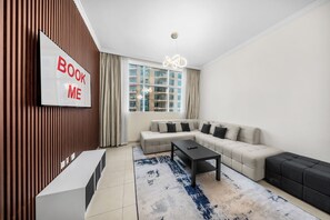 A modern living room with a sectional sofa, a coffee table, a TV mounted on a wooden wall, and a large window with curtains. - StayShort - Elegant 1-Bedroom Stay in Downtown Dubai, Steps from Dubai Mall (Business Bay)