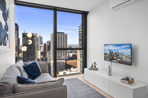A modern living room with a large window, a flat-screen TV, and a sofa with cushions. - High-Rise 1-Bed Apartment in Melbourne CBD (Melbourne)