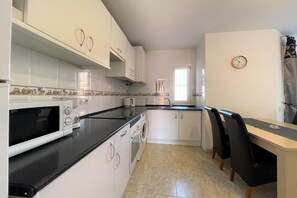 A compact kitchen with white cabinets, a black countertop, a washing machine, and a dining area with chairs. - Coastal Family Apartment in Roquetas de Mar (Roquetas de Mar)