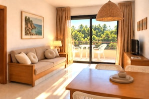 A room with a sofa, a dining table, chairs, a TV, a lamp, and a view of trees through the window. - Coastal Apartment in Pals for 2 (Pals)