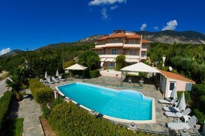 Un hotel con piscina rodeada de sillas de descanso y sombrillas, con montañas al fondo.