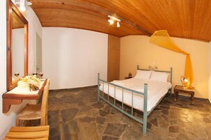 Un dormitorio con suelo de piedra, cama con estructura azul, mesita de noche con lámpara y espejo con un taburete.