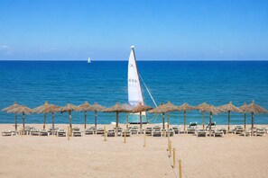 A beach with rows of sun loungers and thatched umbrellas, a sailboat, and calm blue waters. - Beach Apartment with Pool for 5 (Pals)