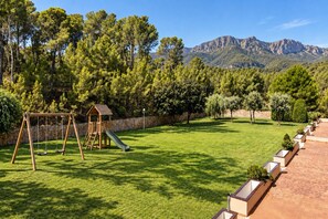 A grassy outdoor area with a wooden playset, including swings and a slide, surrounded by trees and a stone wall. - Beach Apartment with Pool for 5 (Pals)