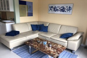 A living room with a beige sectional sofa, a glass coffee table with geometric patterns, and a blue abstract painting on the wall. - Holiday apartment with 1 bedroom (Otterndorf)