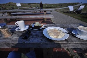Ein Picknicktisch mit Essen und Getränken, eine sitzende Person und ein Blick auf den Strand im Hintergrund.