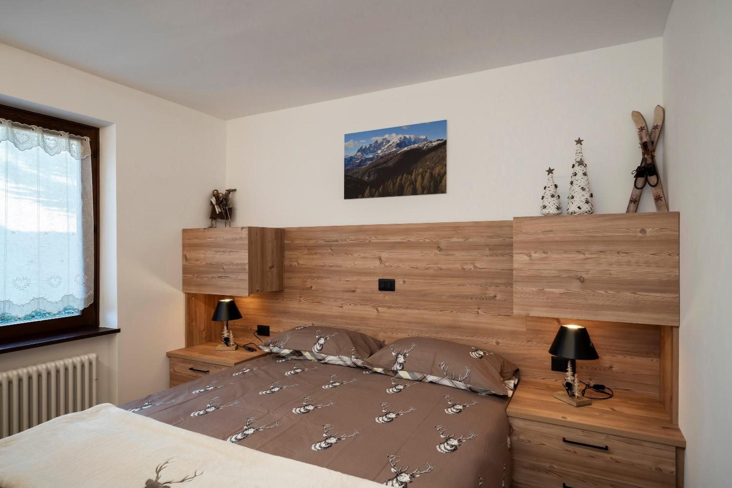 A bedroom with a wooden headboard, a bed with a patterned duvet, bedside lamps, a window with a curtain, and a picture on the wall.