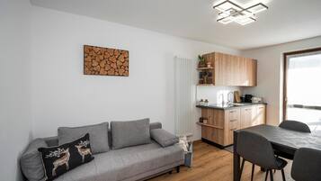 A modern living space with a grey sofa, a dining table with chairs, a kitchen area, and a ceiling light fixture.