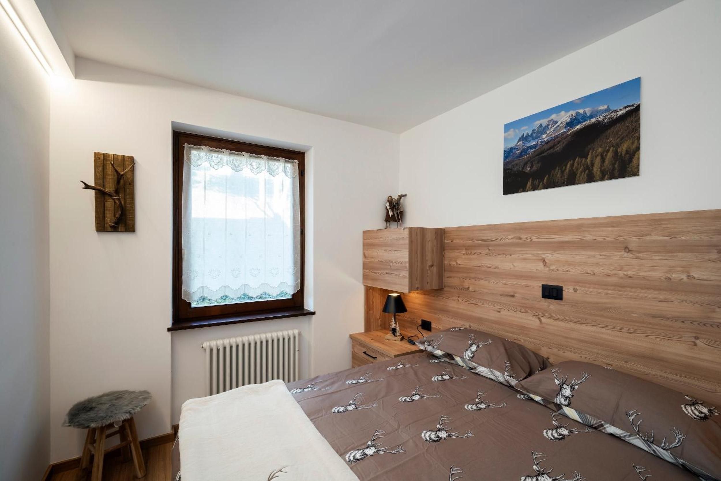 Une chambre avec une tête de lit en bois, une fenêtre ornée de rideaux en dentelle, un radiateur en dessous, un petit tabouret, une table de chevet avec une lampe et un tableau encadré accroché au mur.