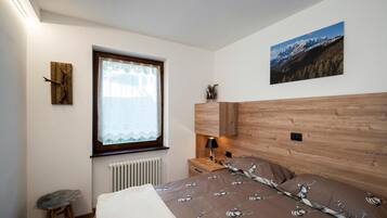 Une chambre avec une tête de lit en bois, une fenêtre ornée de rideaux en dentelle, un radiateur en dessous, un petit tabouret, une table de chevet avec une lampe et un tableau encadré accroché au mur.