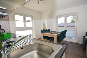 A modern kitchen with a stainless steel sink, a dining table with chairs, and large windows allowing natural light. - Holiday apartment with 1 bedroom (Norden)