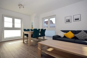A modern living room with a sofa, a dining table with chairs, and a coffee table. There are two windows providing natural light. - Holiday apartment with 1 bedroom (Norden)