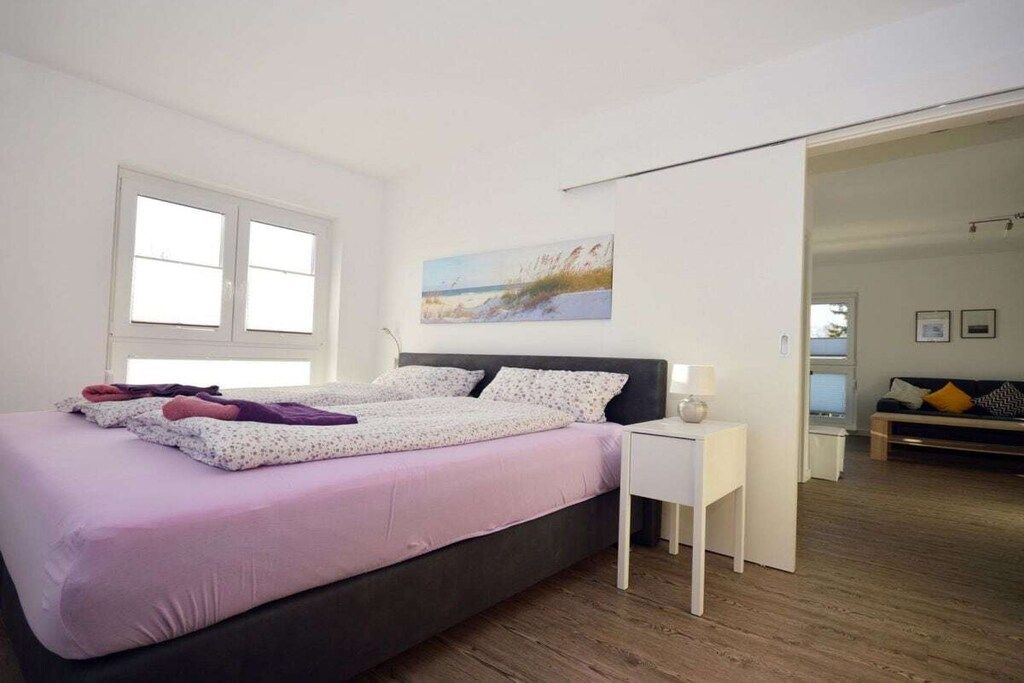 A bedroom with a bed, a nightstand, a painting on the wall, and a view into another room.